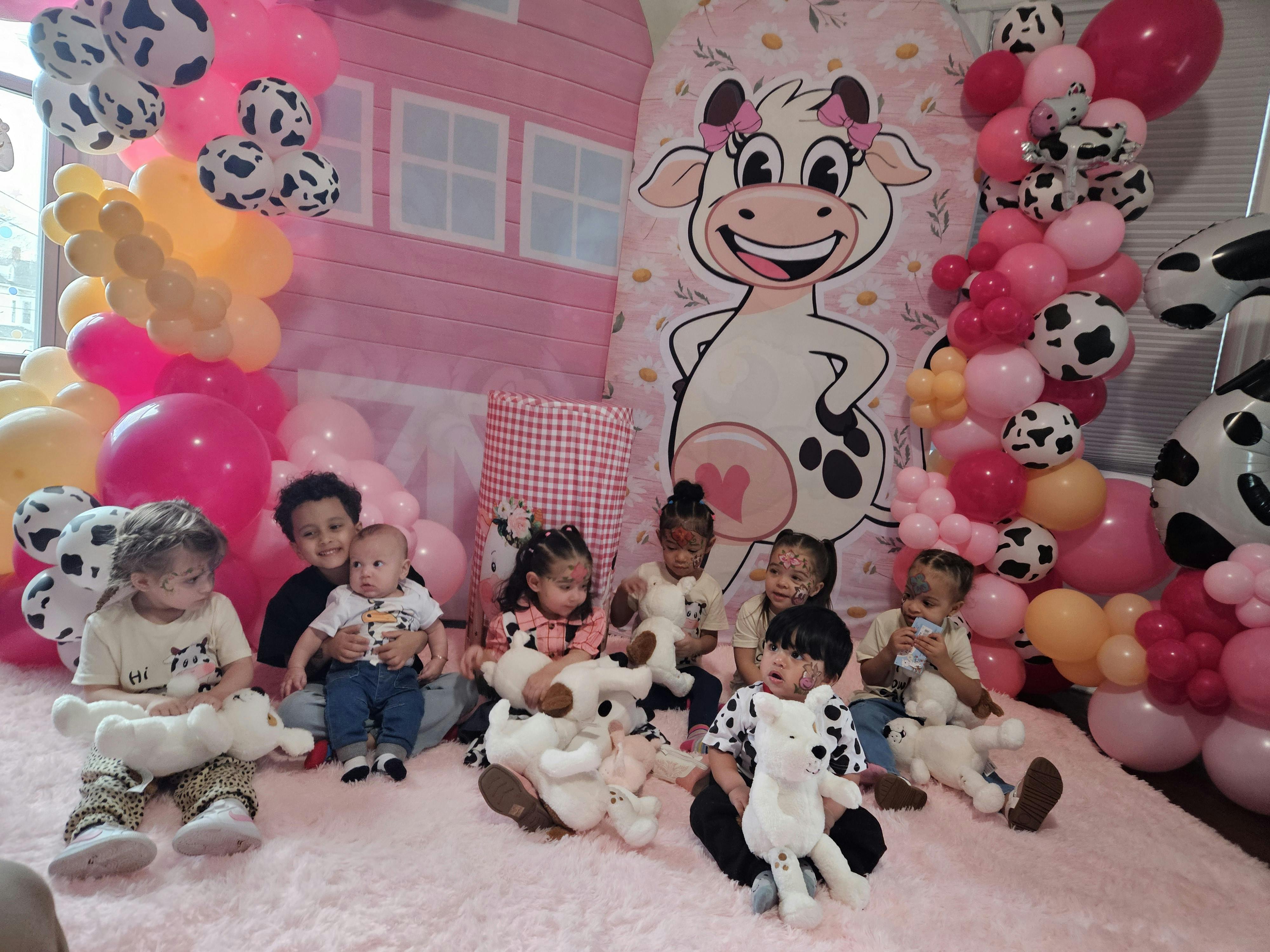 Adults and children sitting on a rug surrounded by balloons and a cow illustration