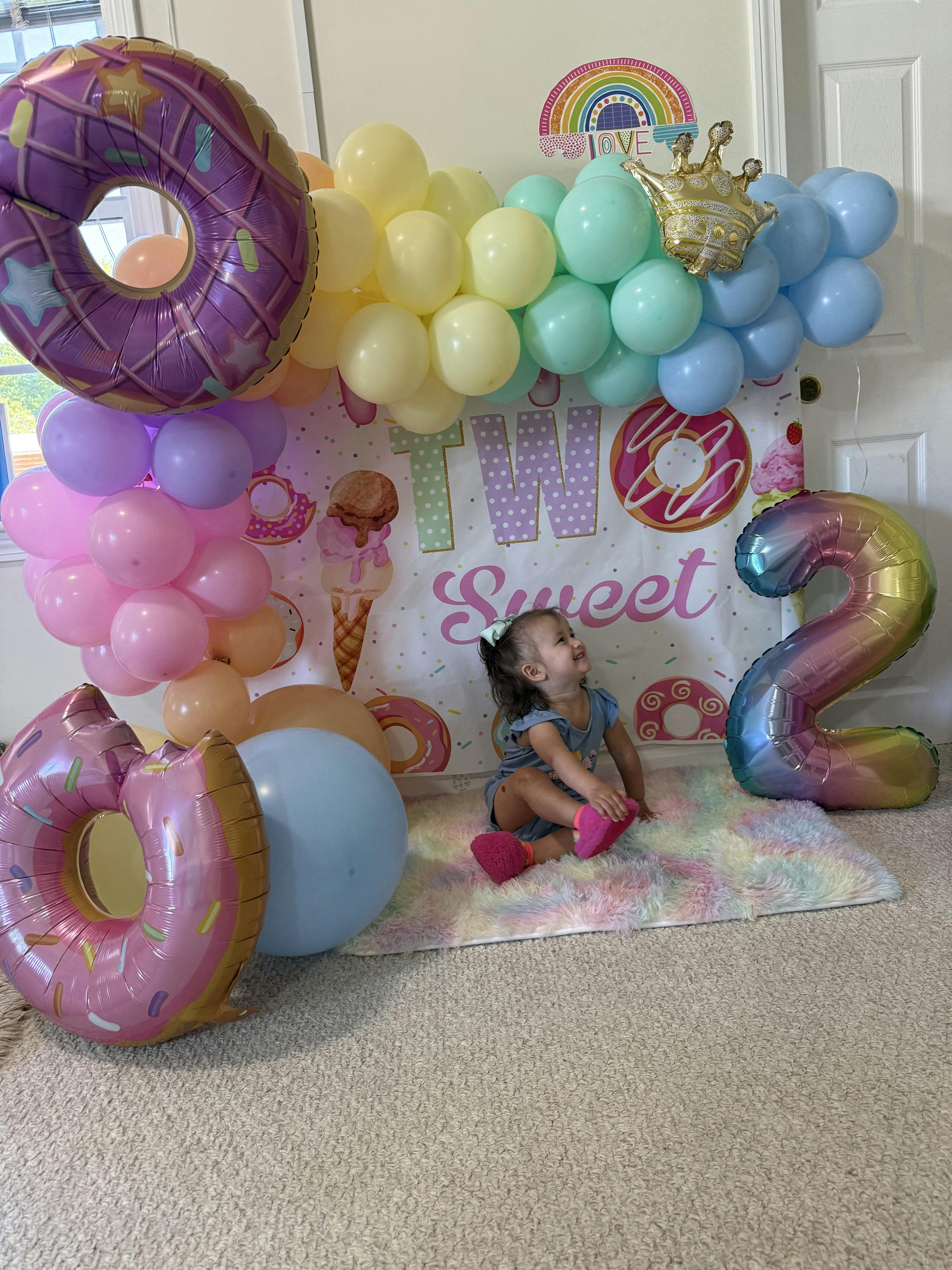 Child sitting on a rainbow rug, surrounded by balloons and donut decorations