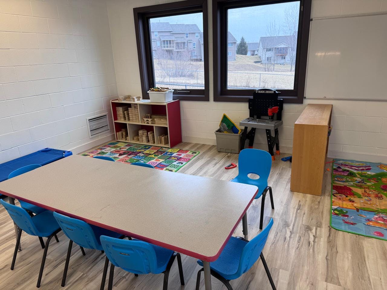 Classroom with a long table and blue chairs, shelves of blocks, and play rugs