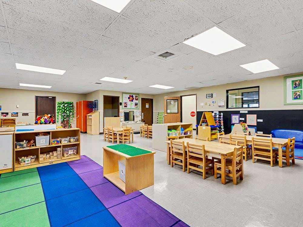 Empty classroom with small tables and chairs, storage shelves, and colorful rug