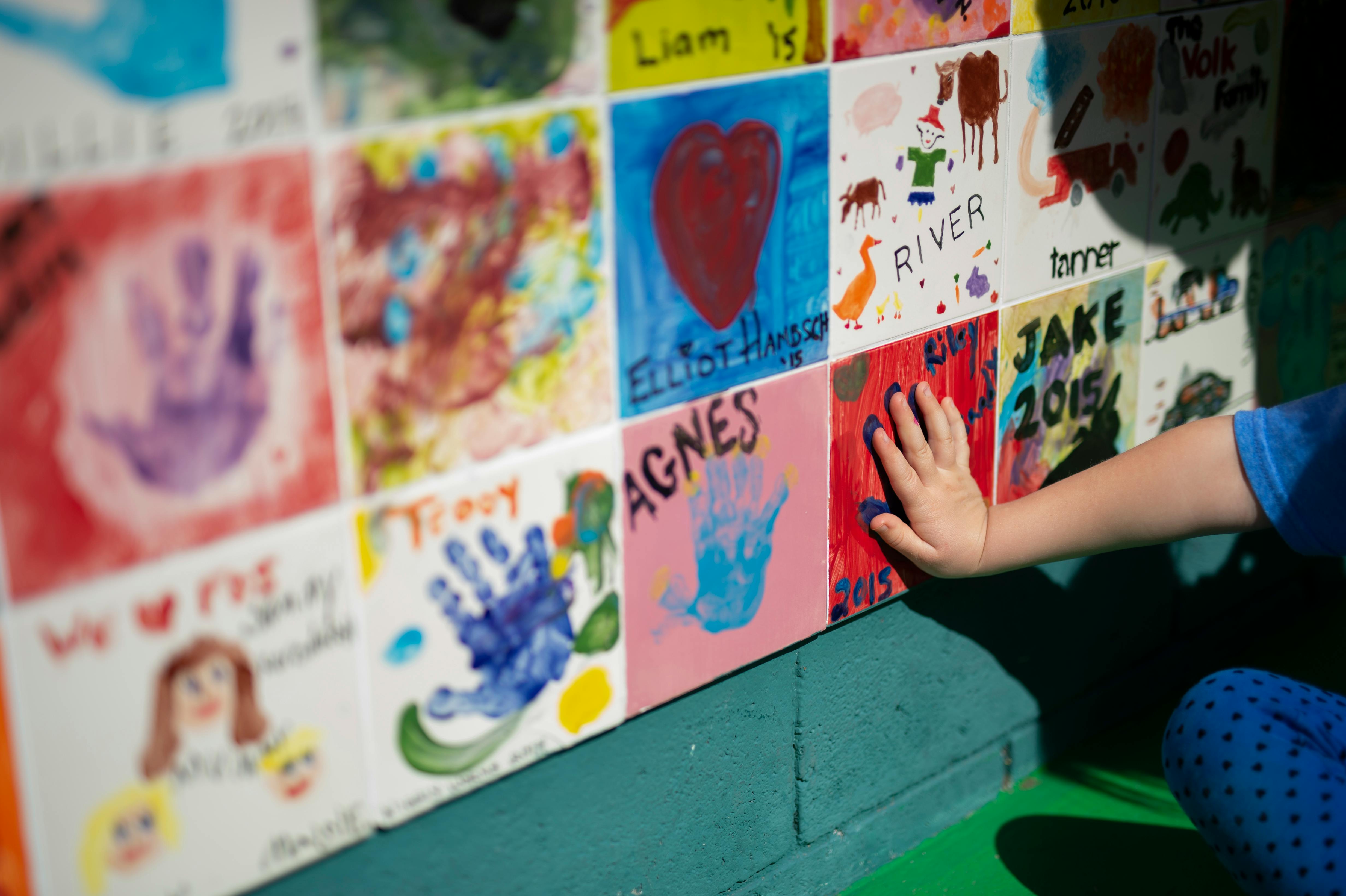 Child's hand touching a wall of colorful art tiles with handprints and names