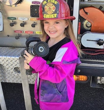 Child in a Jr. Fire Chief helmet holding a fire hose nozzle