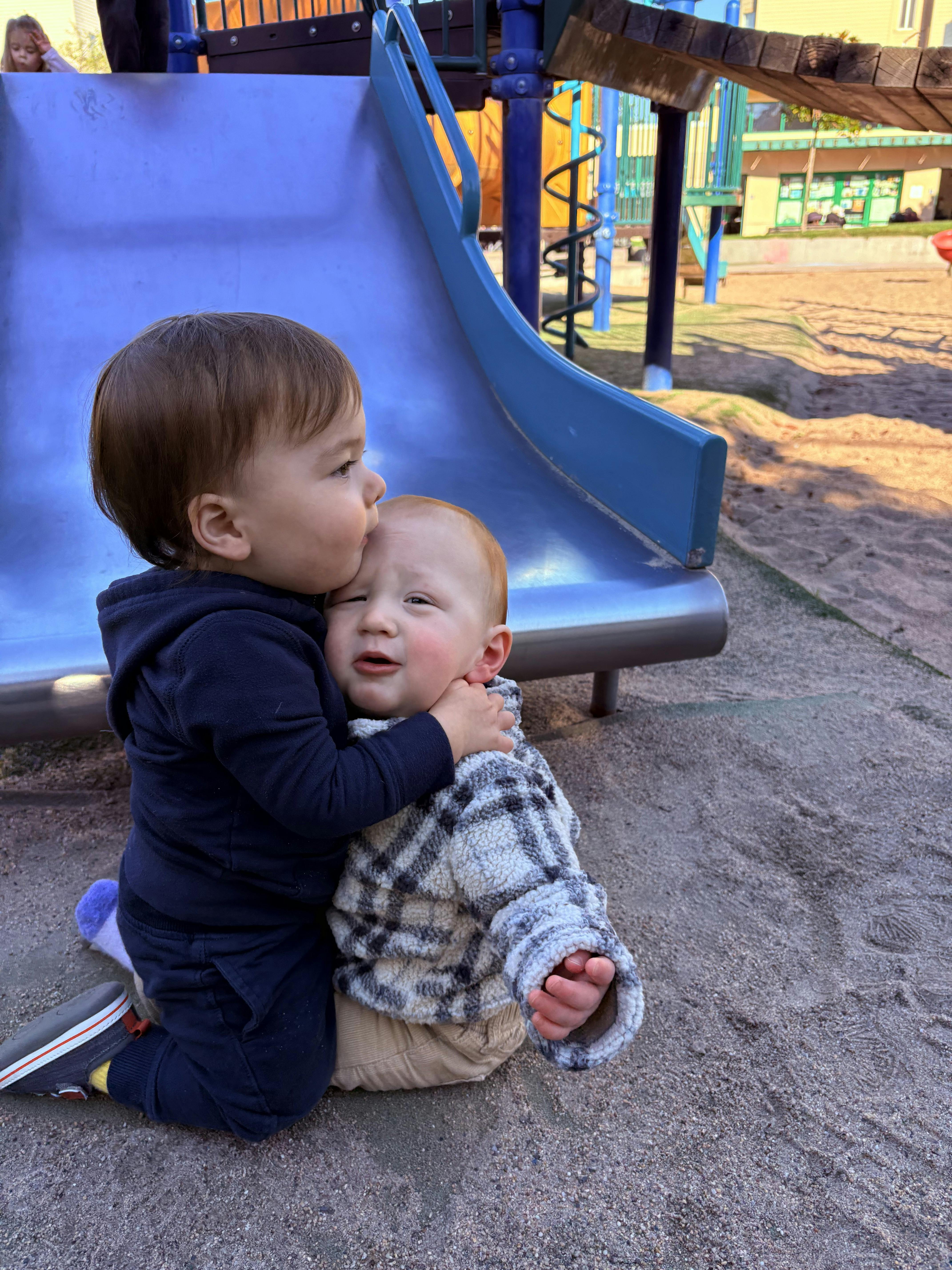 One child kneeling and kissing the forehead of another child on a playground