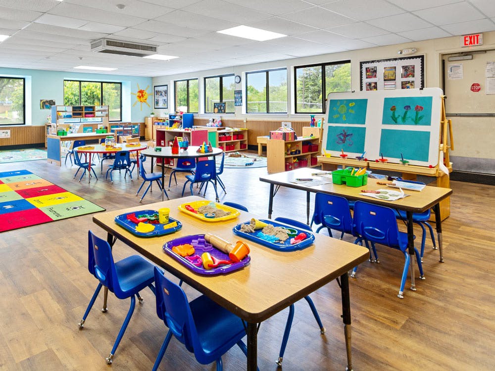 Classroom with small tables and chairs, toys, and an easel with children's drawings