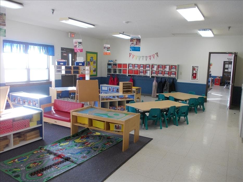 An empty classroom with small tables, chairs, shelving units, and a playmat
