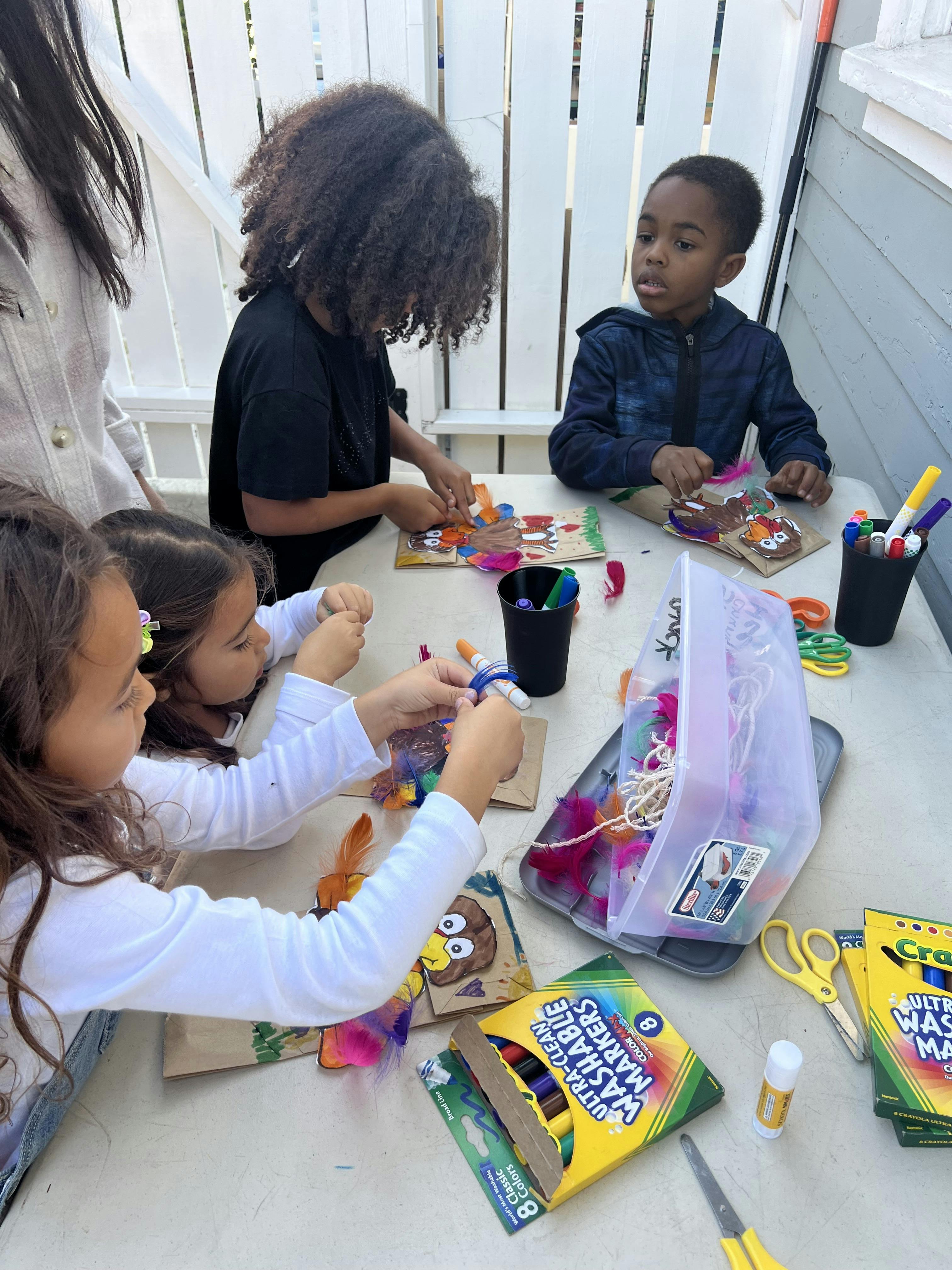 Three children at a table decorating paper bags with feathers and markers