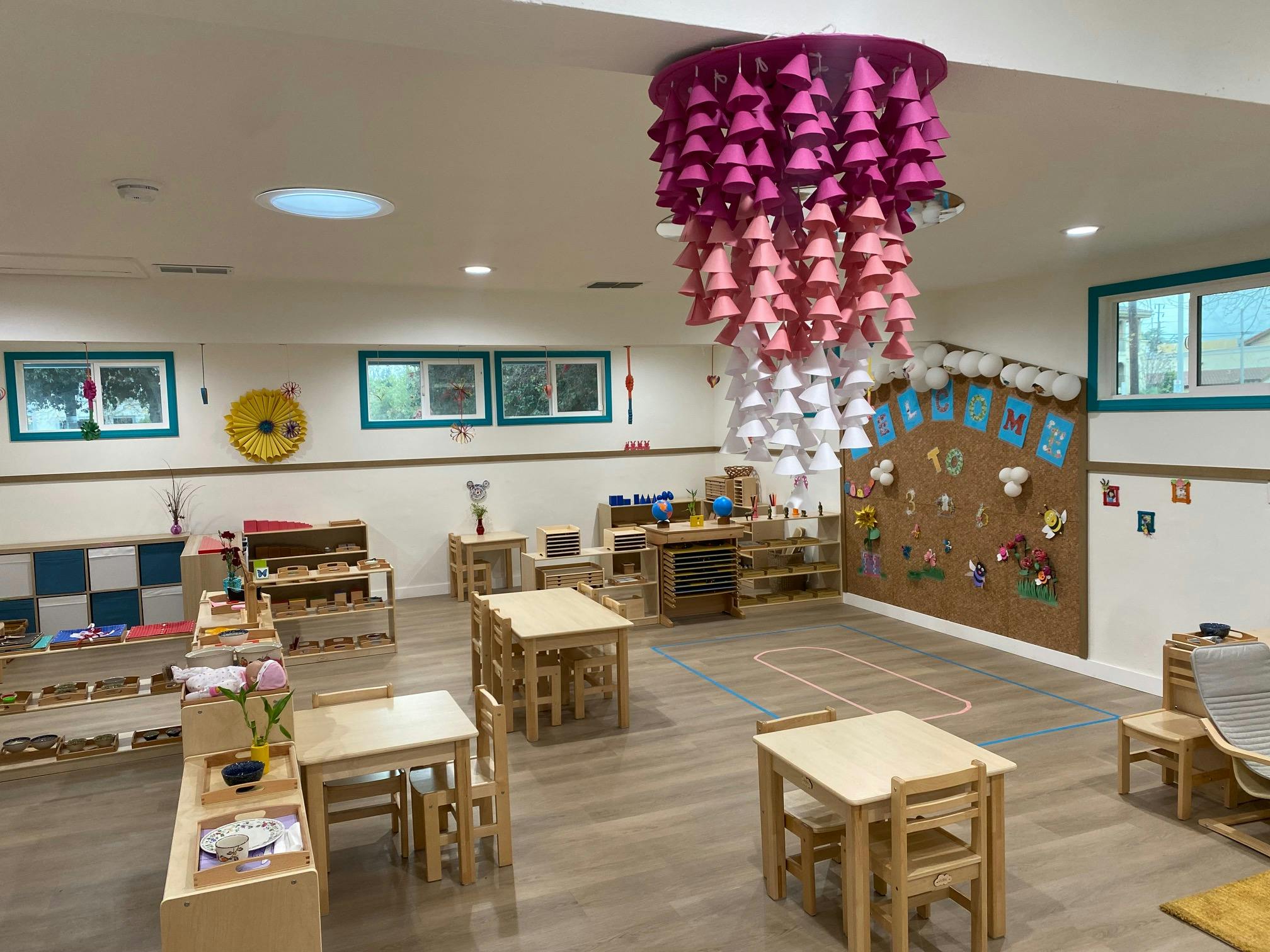 Classroom with small wooden tables and chairs, shelves with learning materials