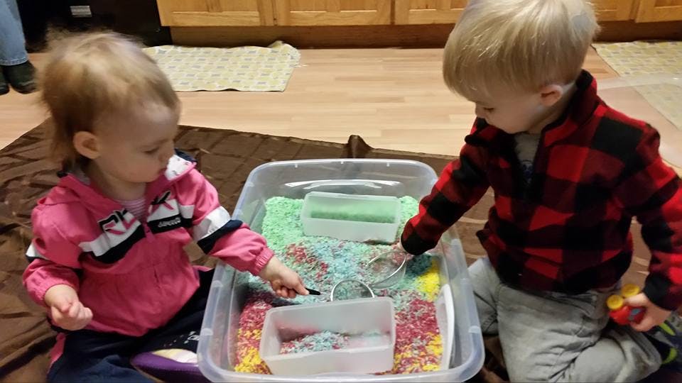 Two children playing with colorful sensory bin materials in a clear bin