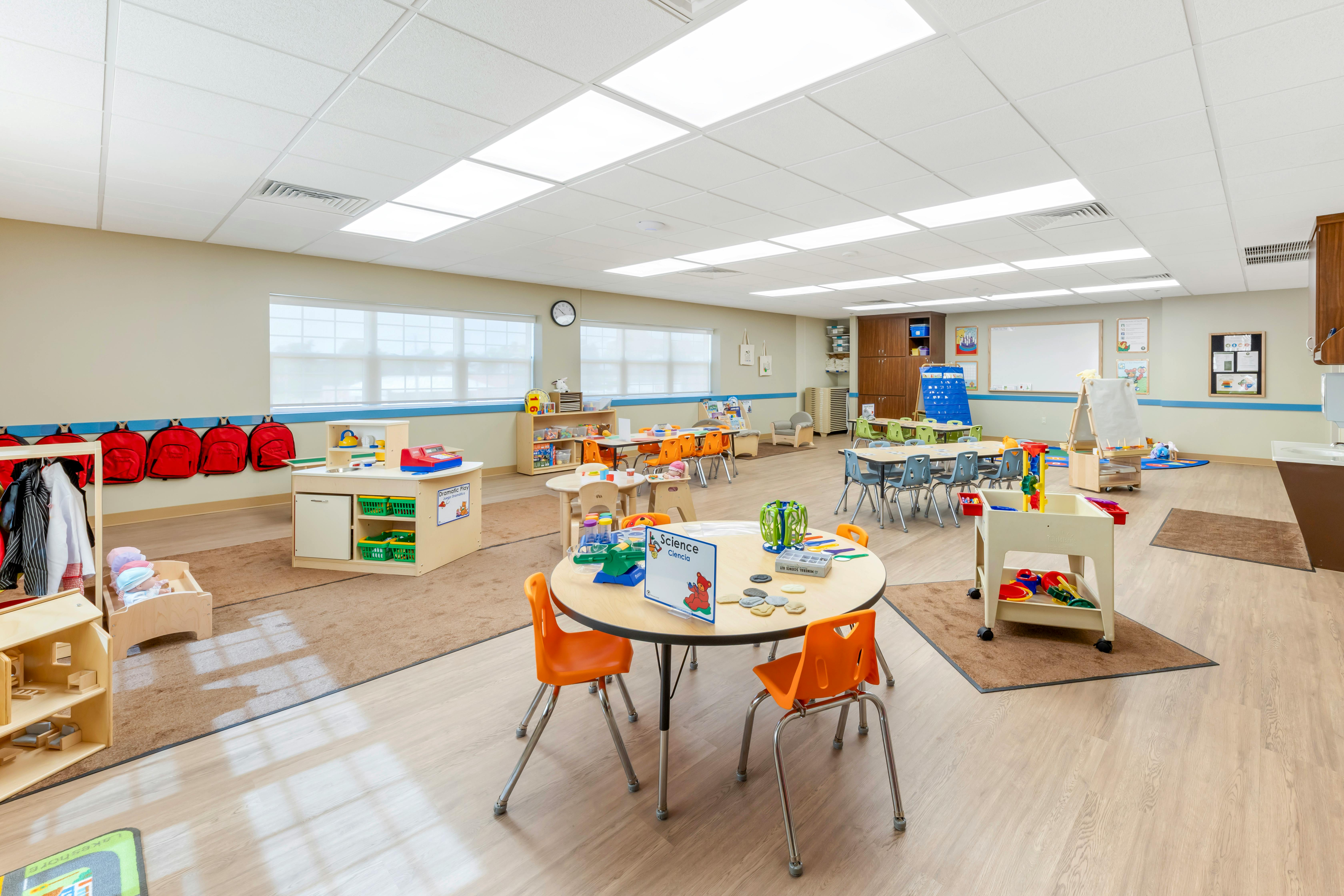 Classroom with child-sized tables and chairs, play kitchen, and red backpacks