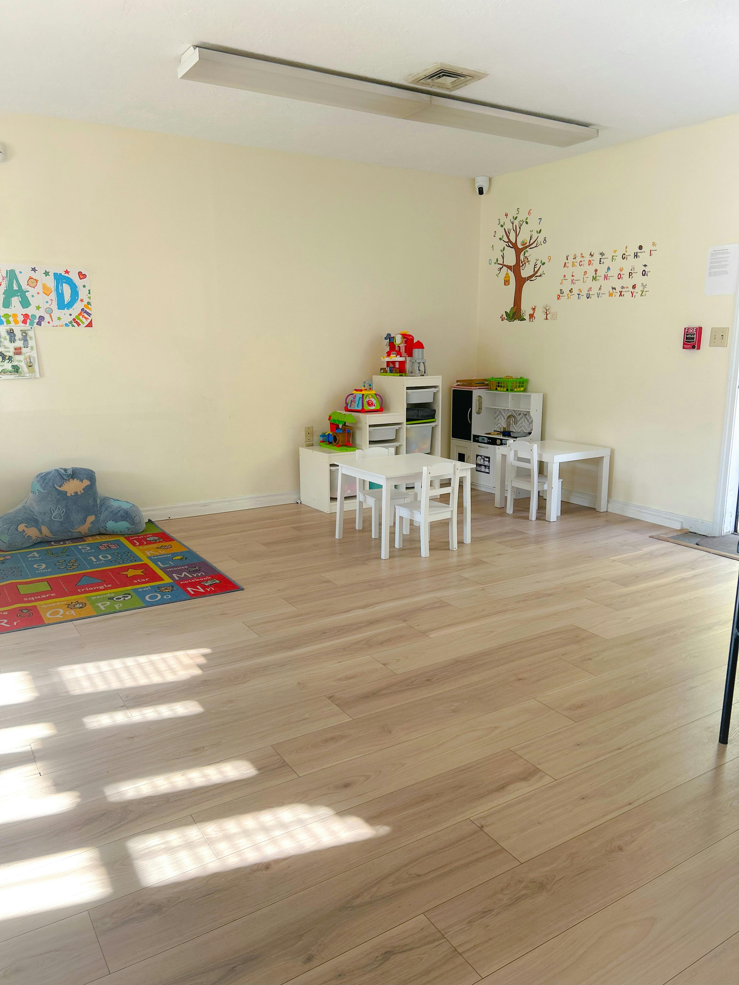Empty classroom with small tables and chairs, a colorful rug, and wall decorations