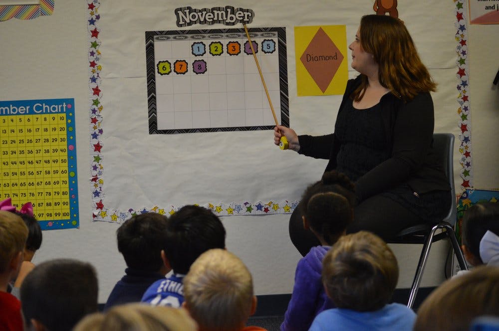 Person teaching children in a classroom, pointing to a calendar with a stick