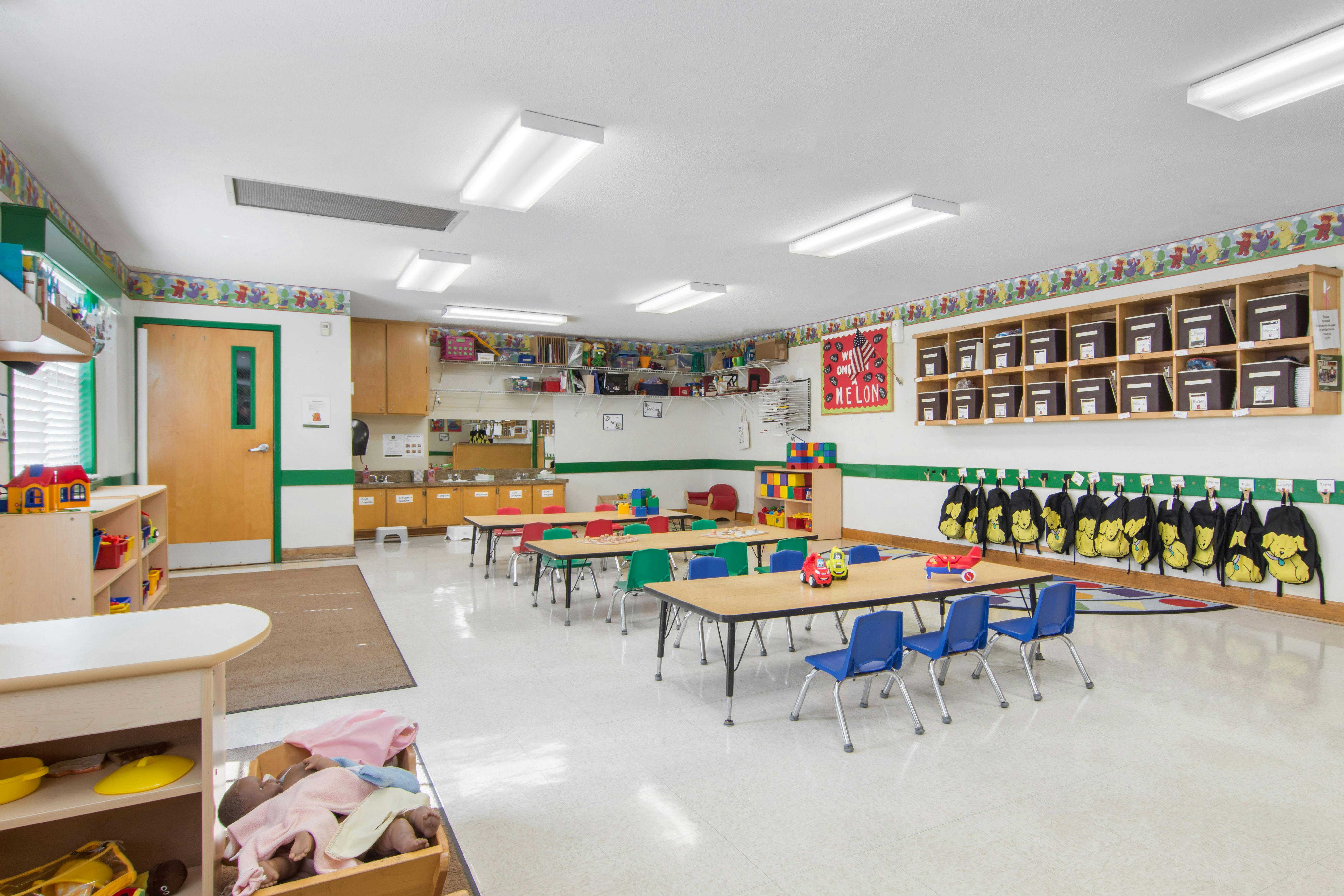 Classroom with small tables and chairs, shelves, and cubbies with yellow backpacks