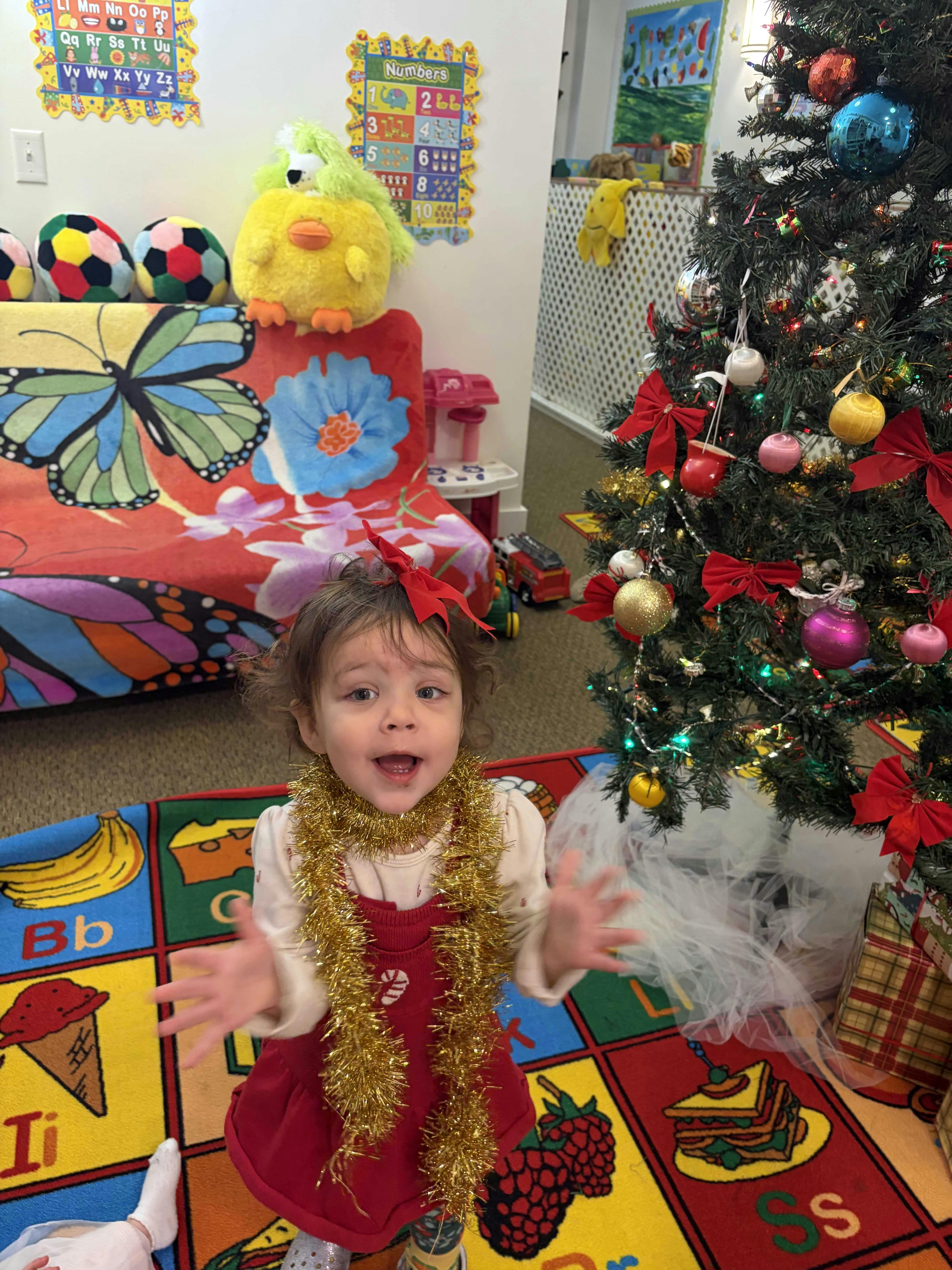 Child wearing gold tinsel standing on an alphabet rug near a Christmas tree