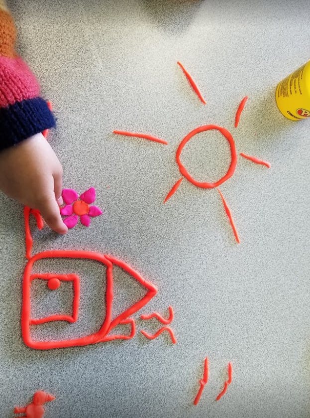 Child's hand placing a pink flower on a red play-doh house with a sun