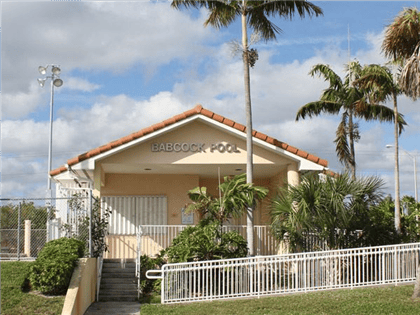 Building with "BABCOCK POOL" sign, palm trees, and white fence