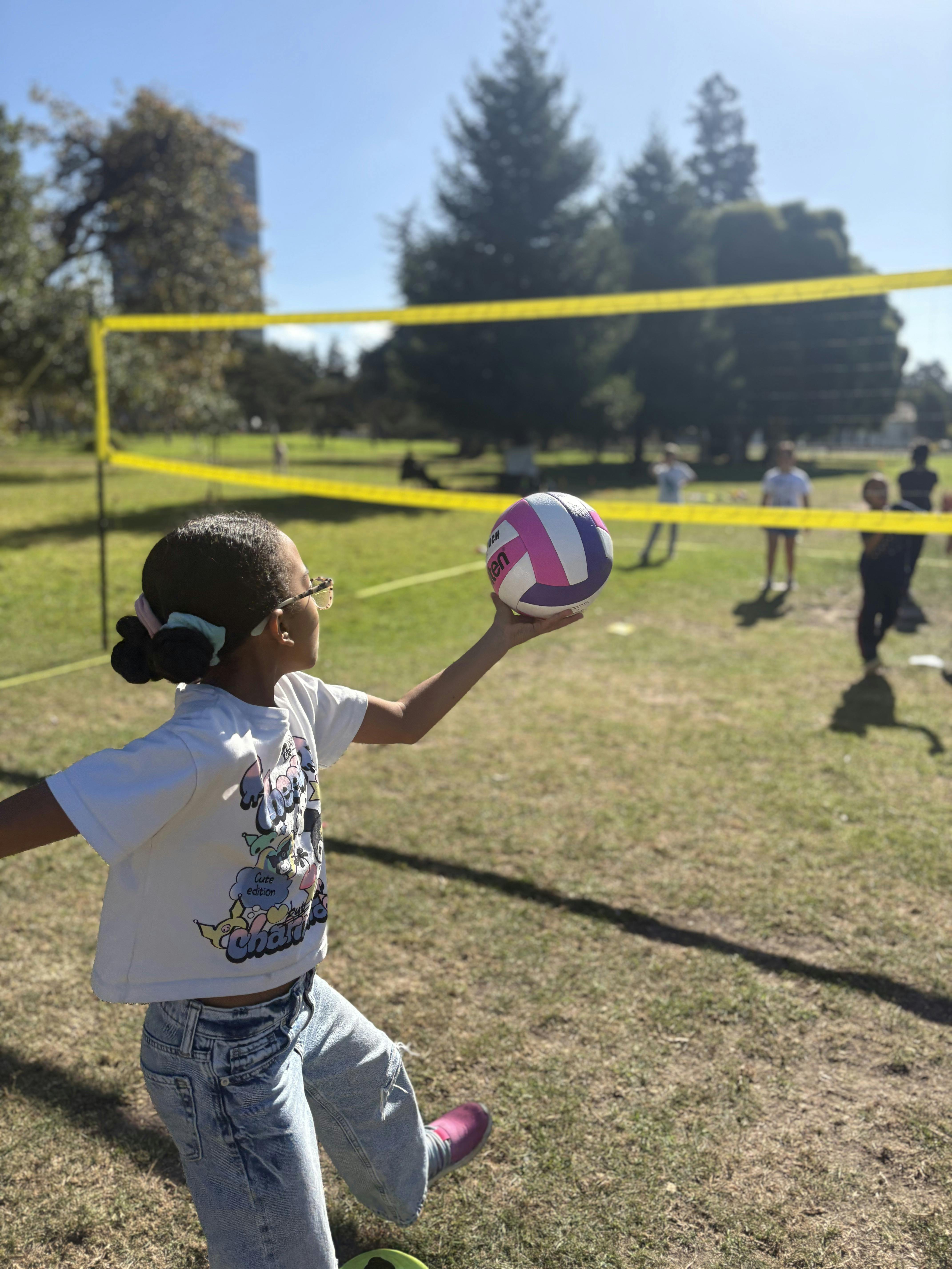 Child holding a volleyball ready to serve with a net and other children in the background
