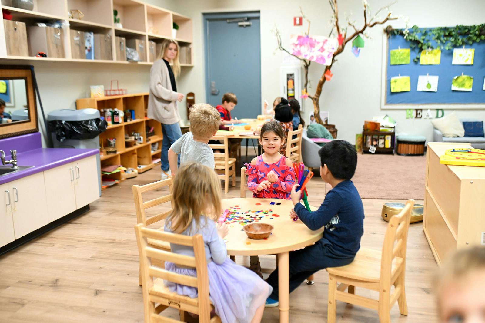 Children sitting at tables in a classroom, some playing with toys