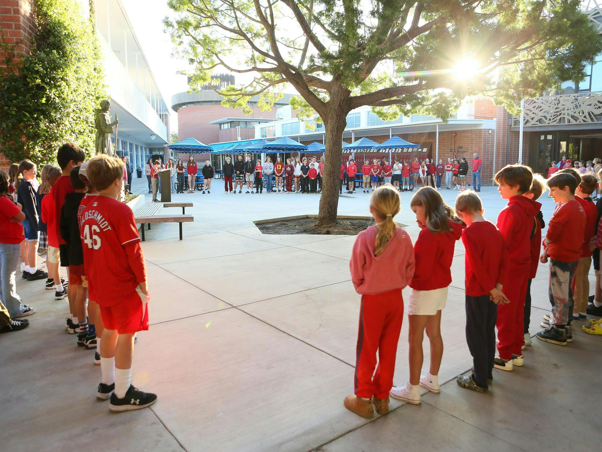Children gathered in an outdoor courtyard with a person speaking at a podium
