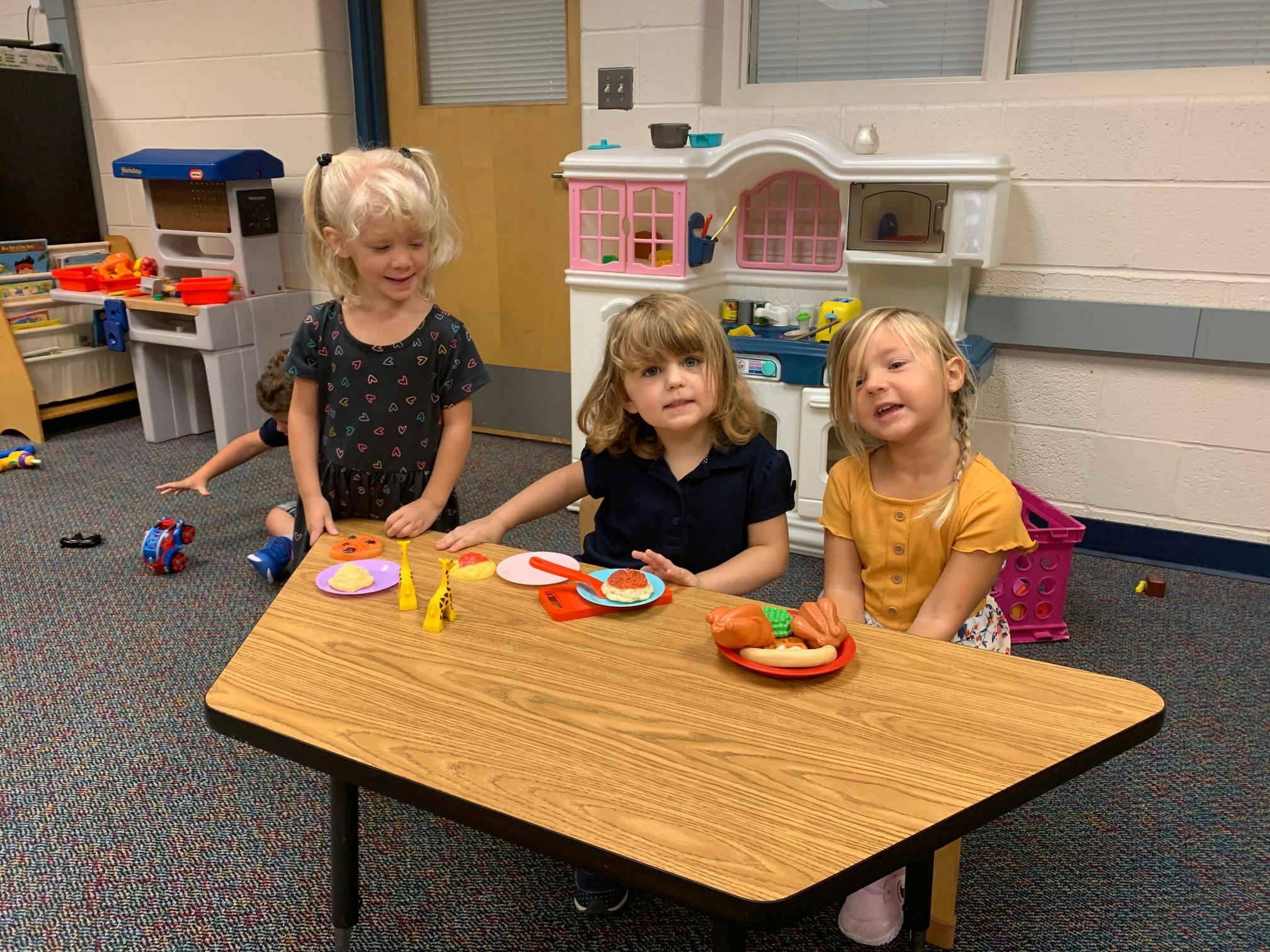 Three children playing with toy food at a small table in a classroom