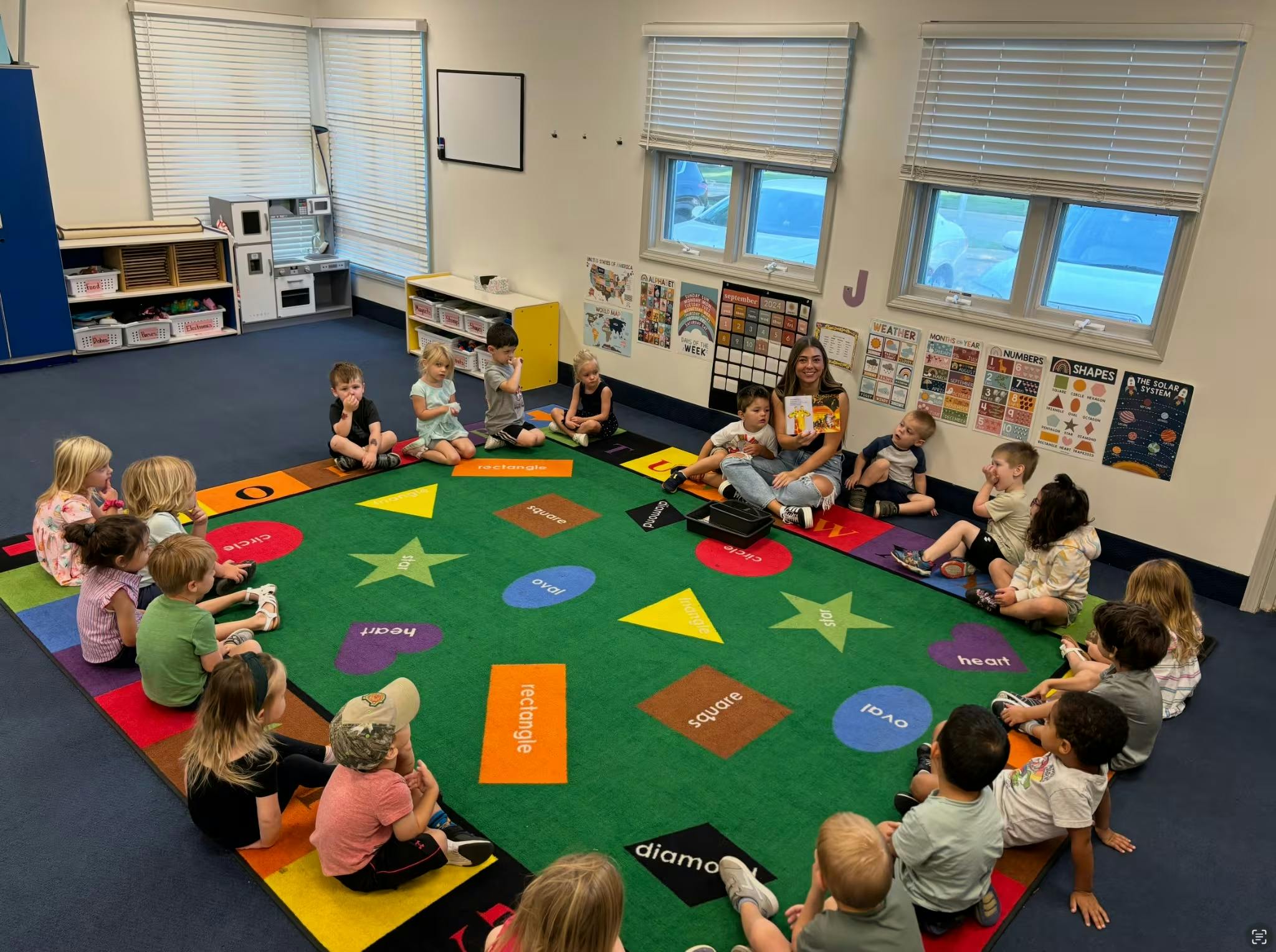 adult reading a book to a group of children seated on a colorful rug in a classroom