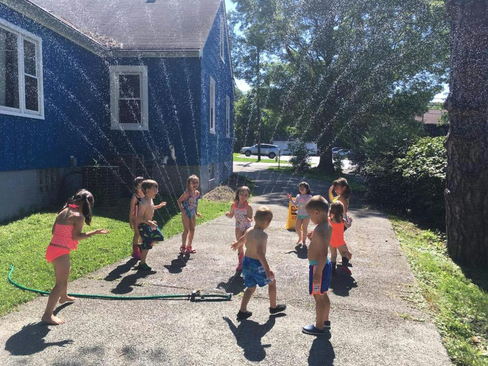 Children playing in a sprinkler on a paved surface next to a house