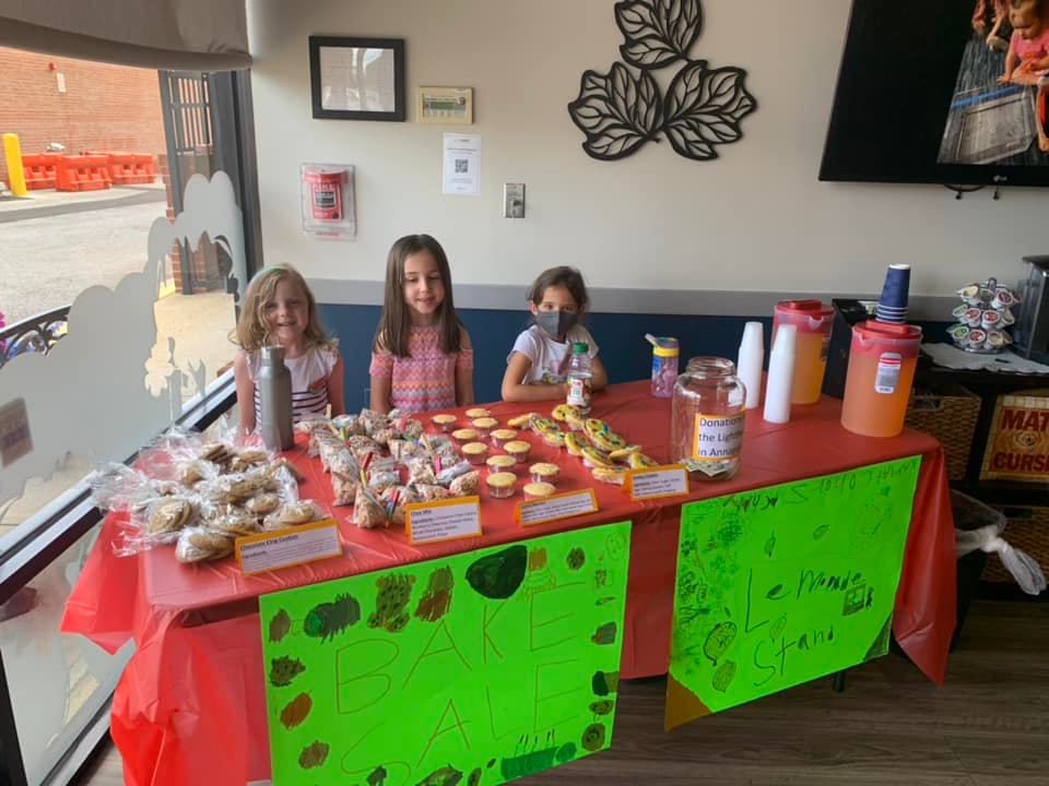 Three children behind a table with baked goods and drinks
