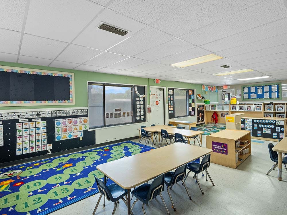 An empty classroom with small tables and chairs, a number rug, and teaching aids