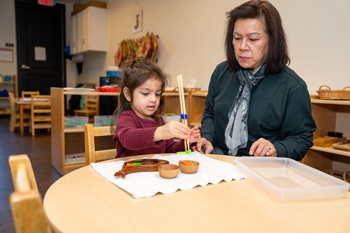 Adult and child using chopsticks to move small objects in a classroom