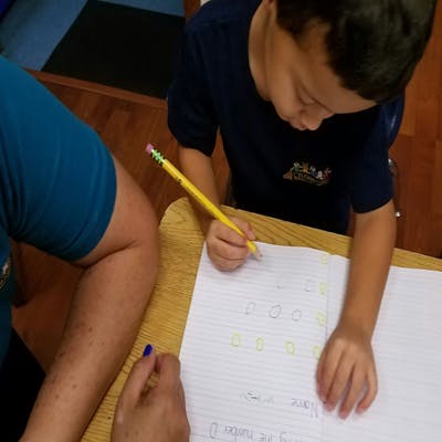 Child writing on paper with a pencil, person's arm next to them