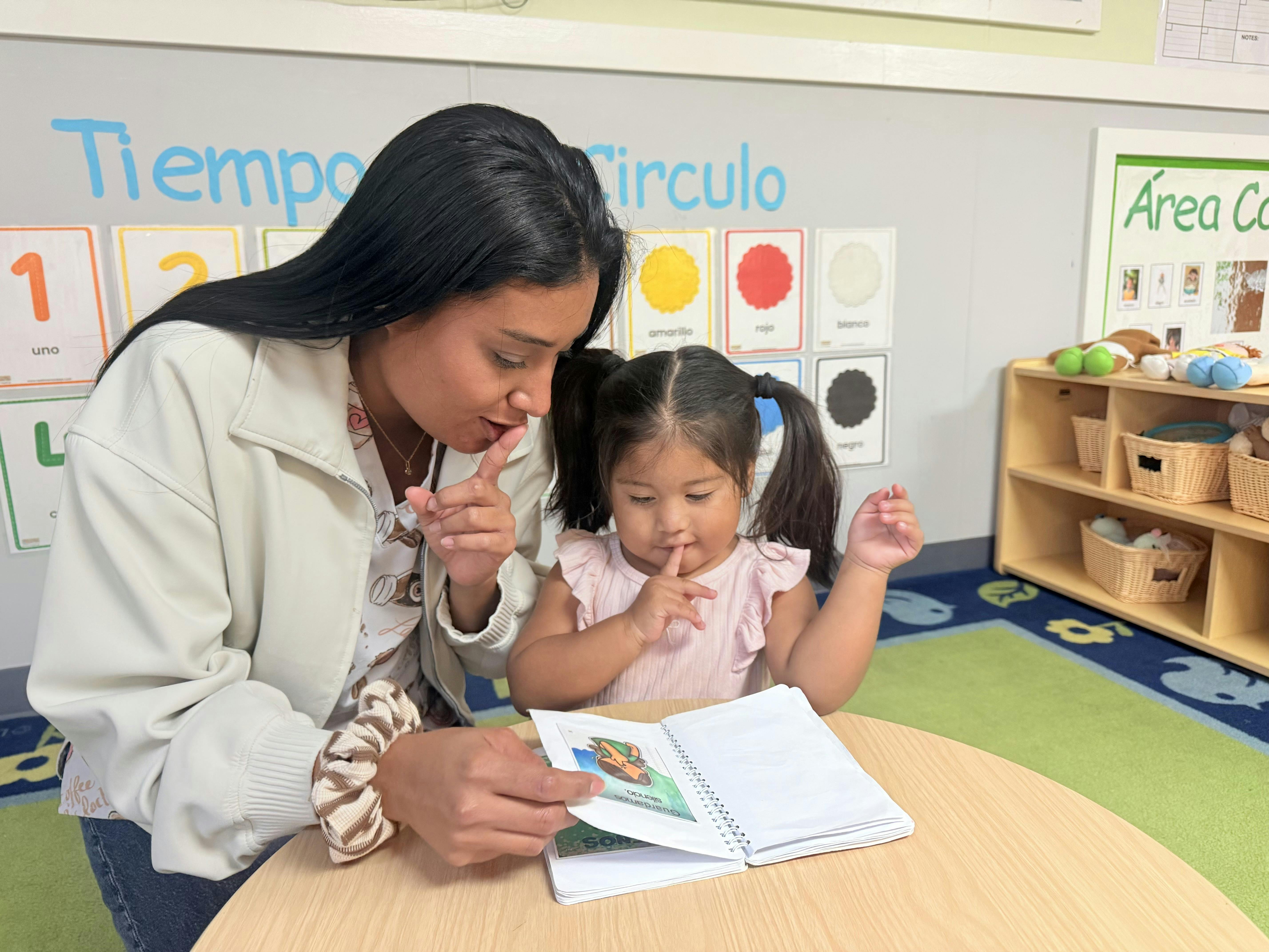 Adult and child in a classroom holding a finger to their lips while reading
