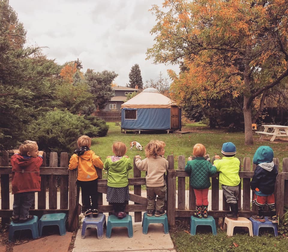 Children standing on stools looking over a fence at a yurt