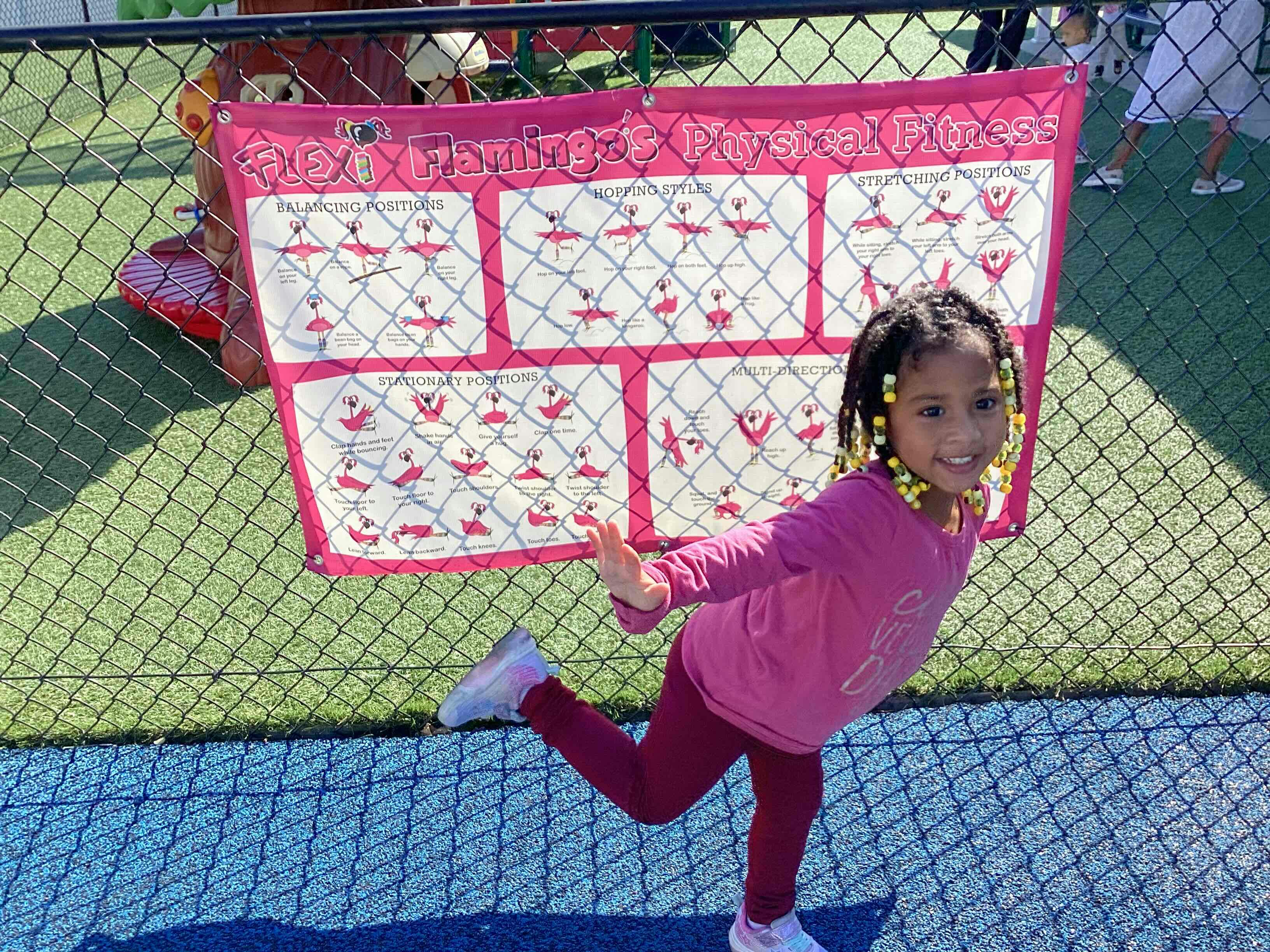 Child balancing on one leg in front of a fence with a sign
