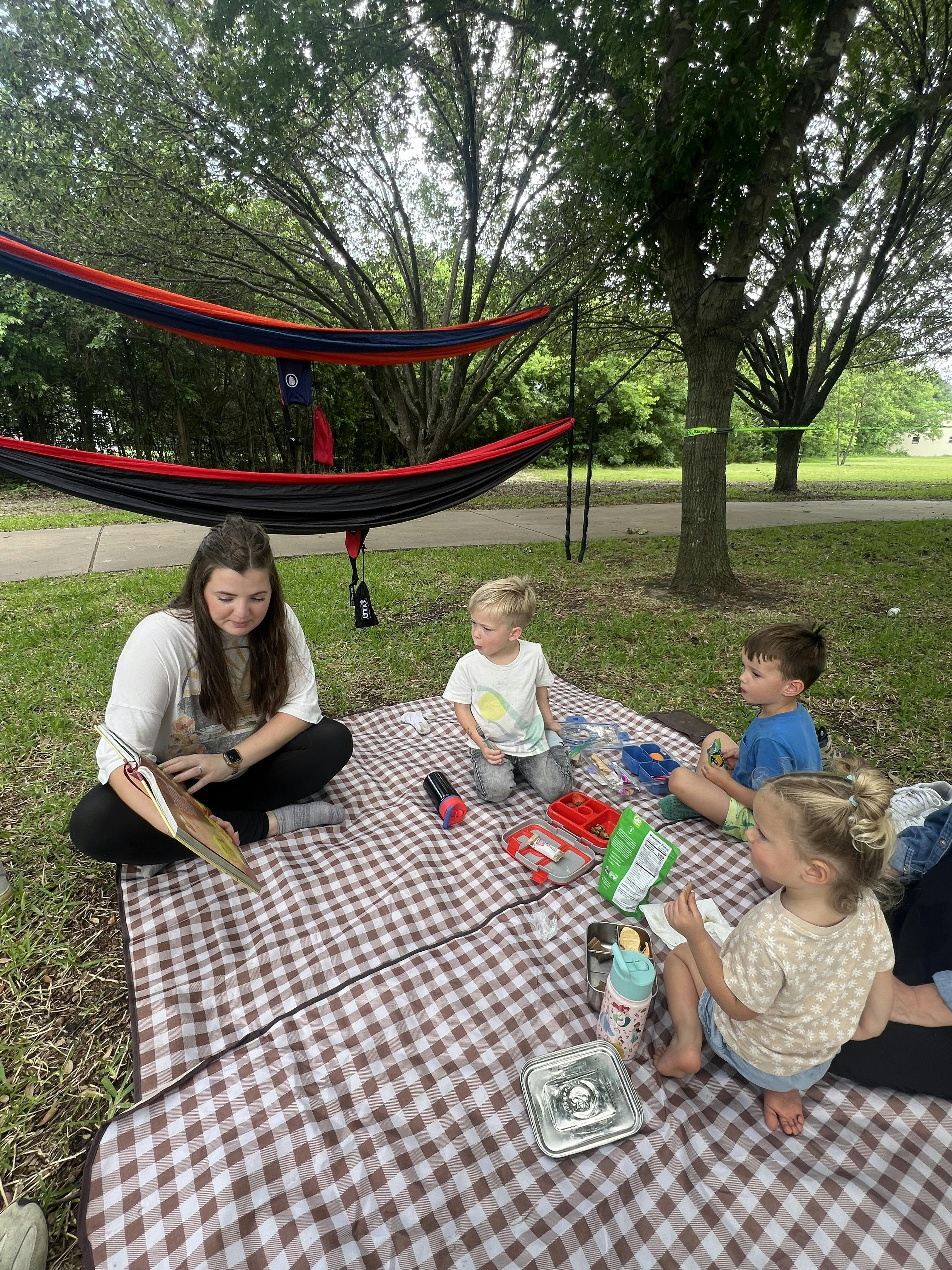 Person reading to three children sitting on a picnic blanket outdoors