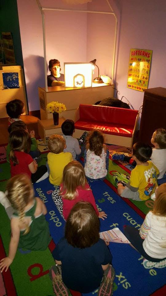 Children sitting on a rug watching a person behind a lit screen