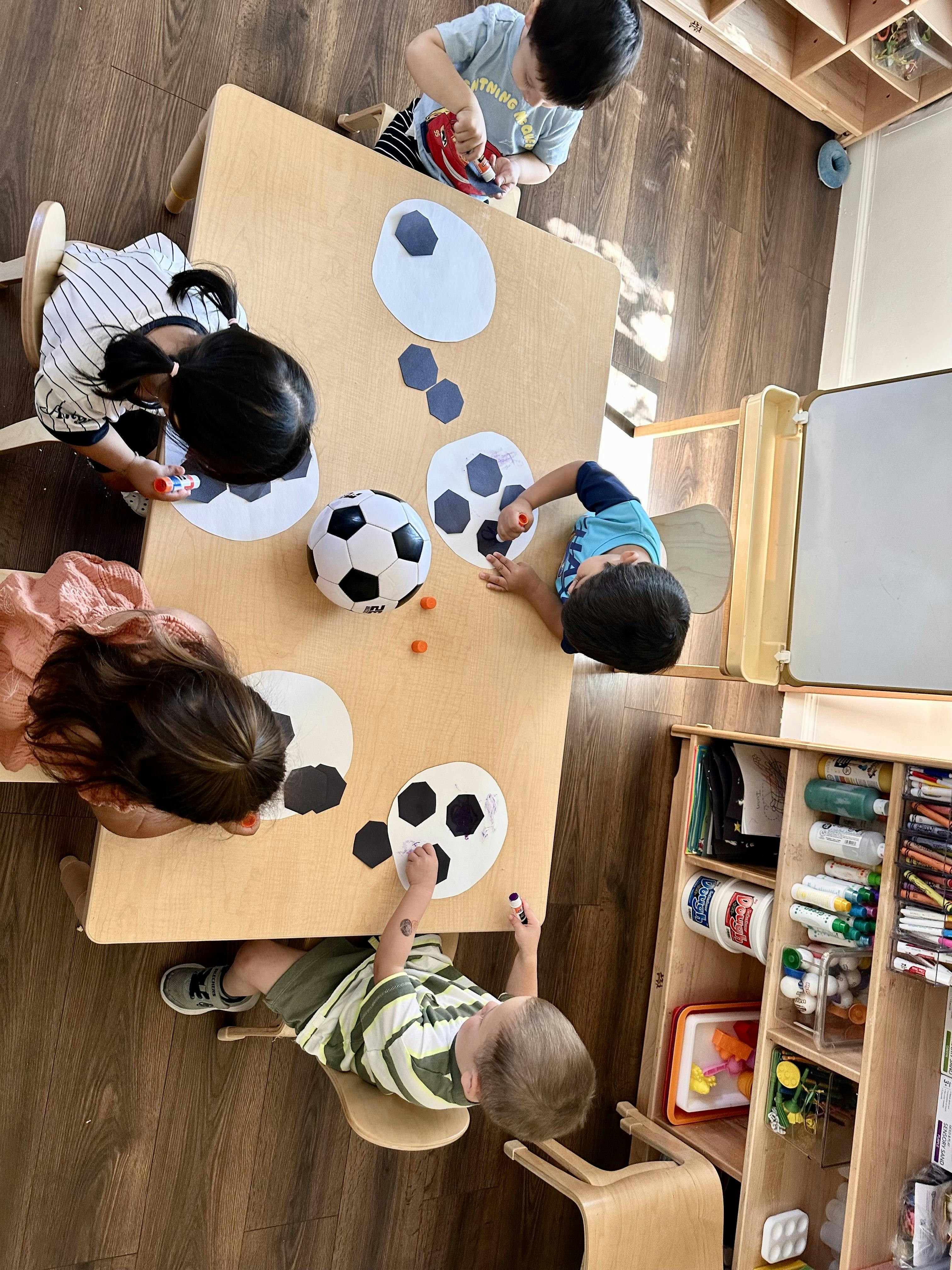Children at a table gluing paper shapes onto soccer ball cutouts