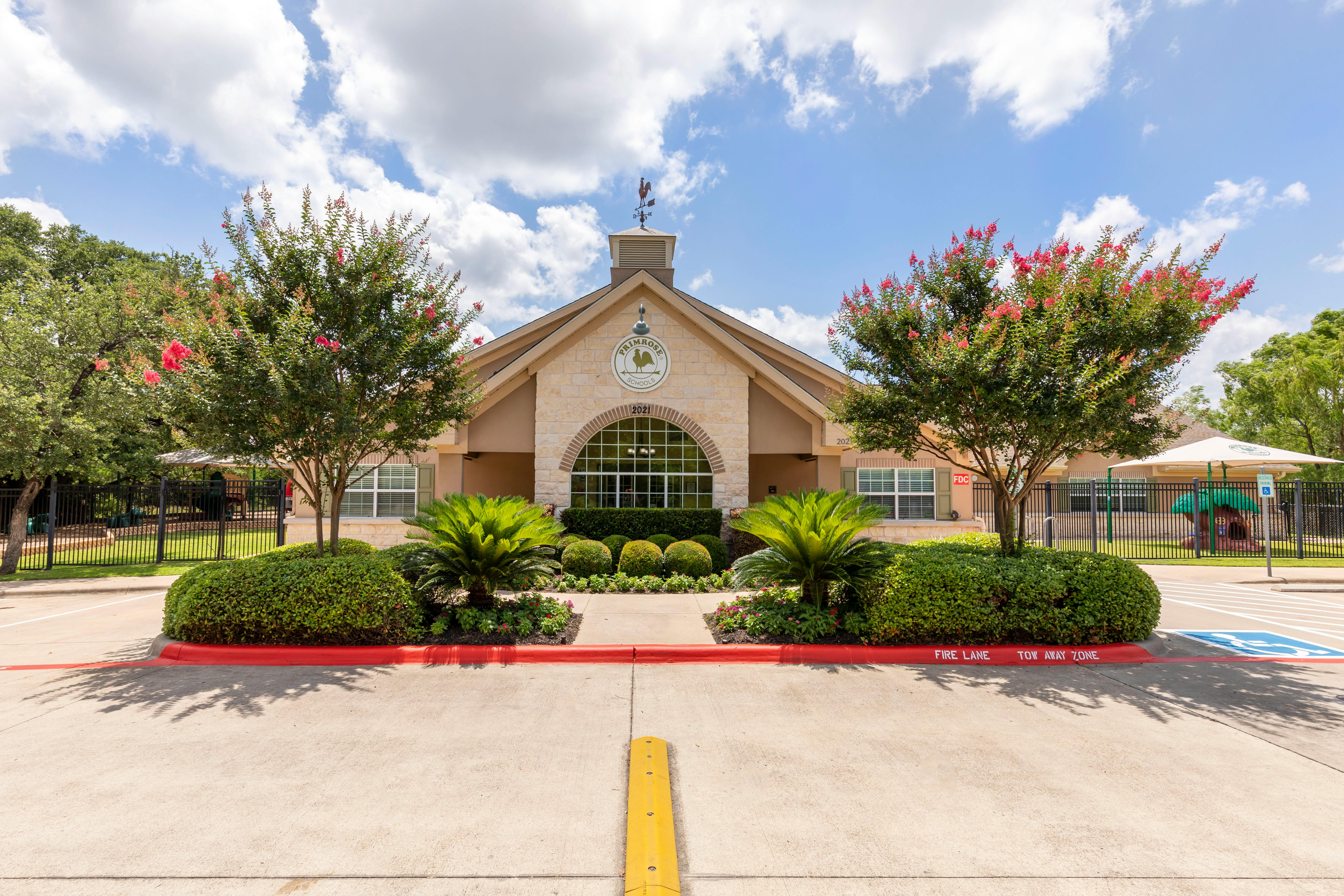 Front exterior of a Primrose Schools building with a courtyard, landscaping, and a fenced playground