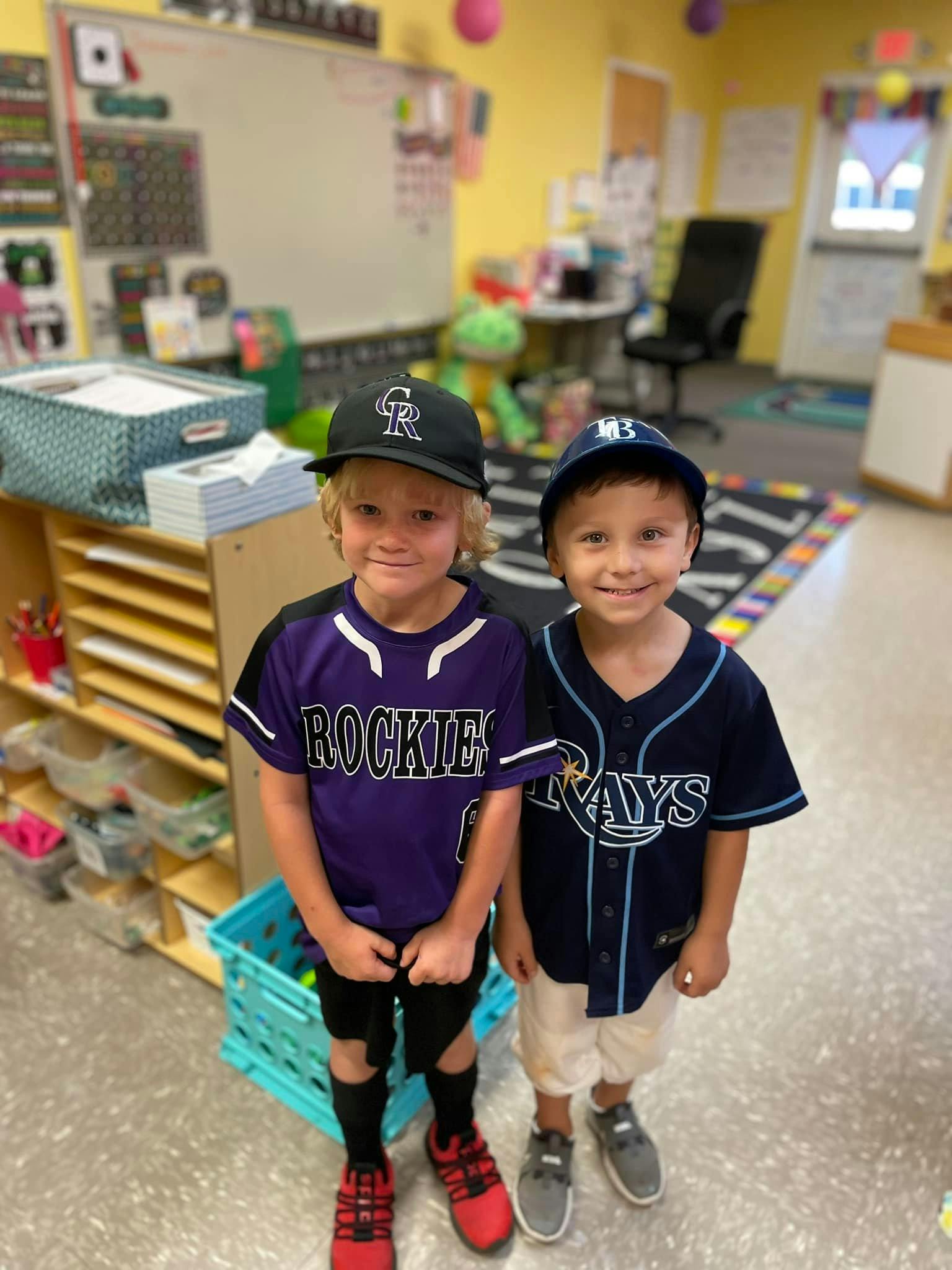 Two children in baseball jerseys and caps standing in a classroom