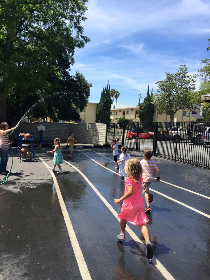 Person spraying water from a hose at children running on a wet paved surface