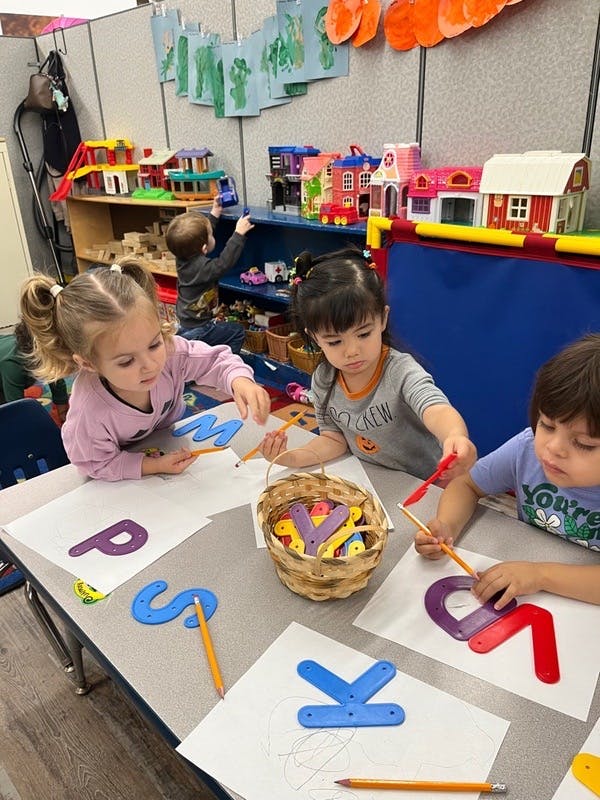 Three children tracing letters at a table in a classroom