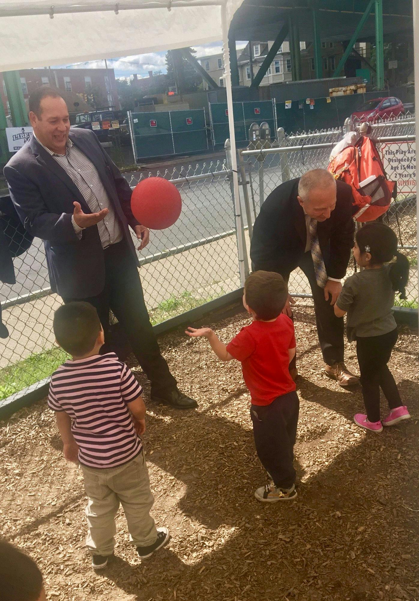 Two men interacting with three children, one man holding a red ball