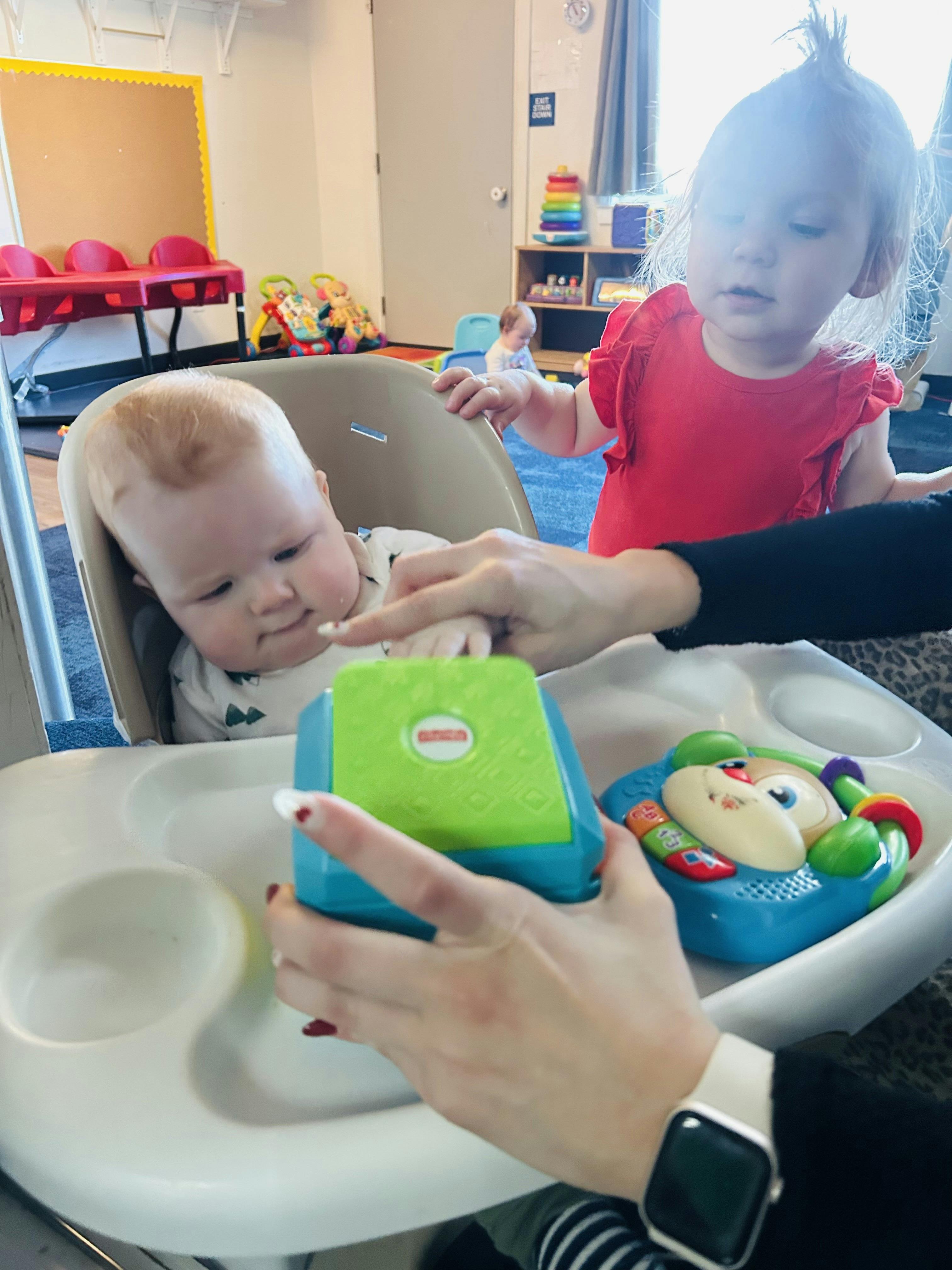 Child in high chair reaching for toy, adult hands holding toy