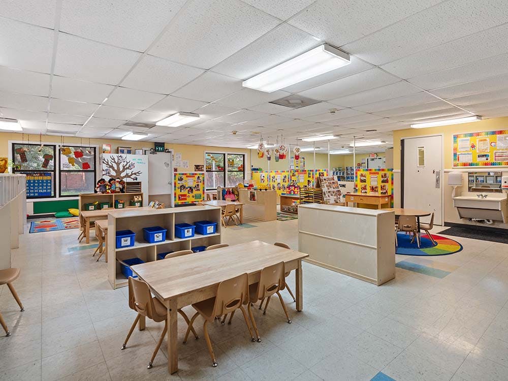 Empty classroom with small tables and chairs, shelves with bins, and windows