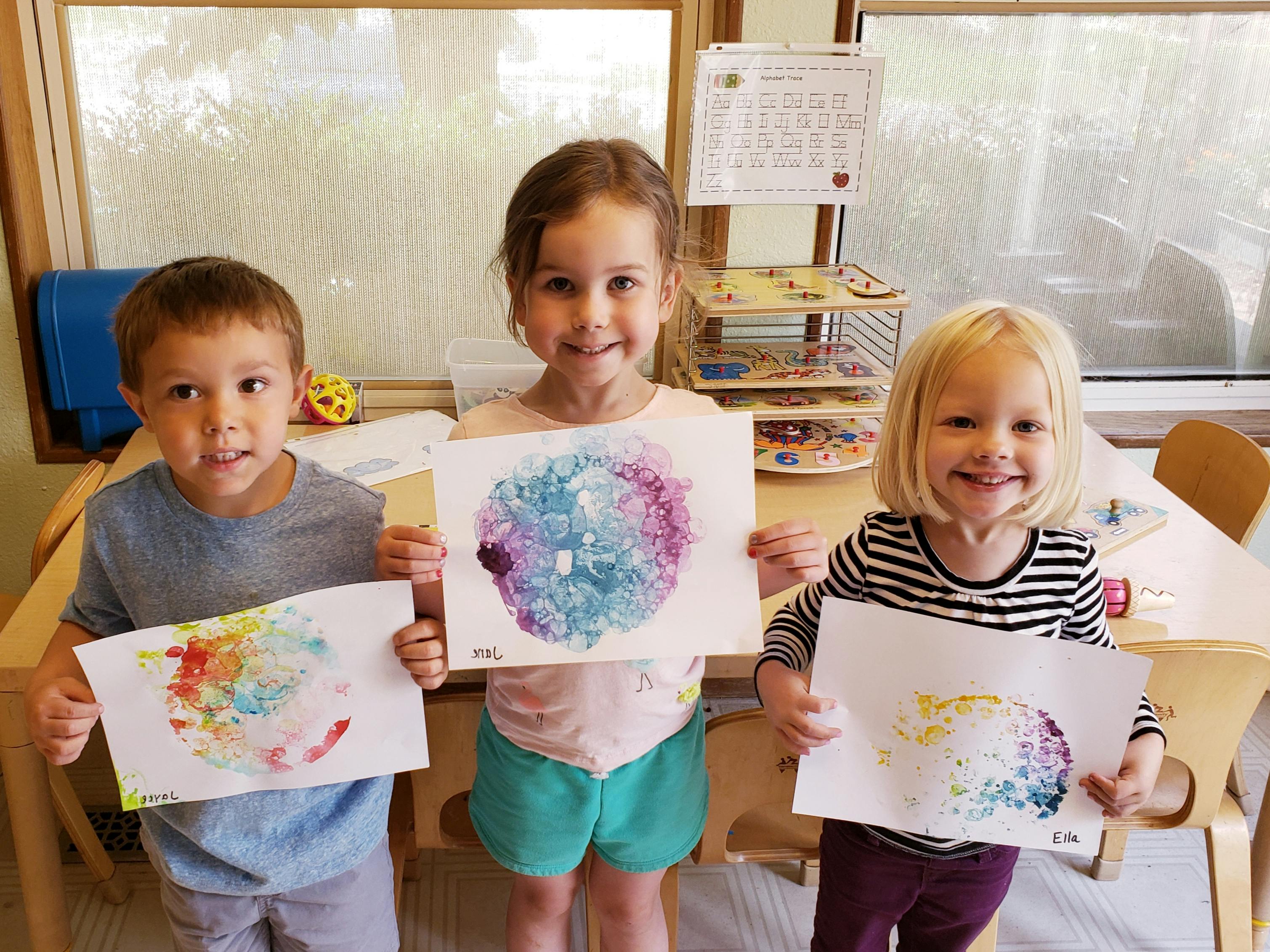 Three children holding up their colorful paintings in-progress paintings