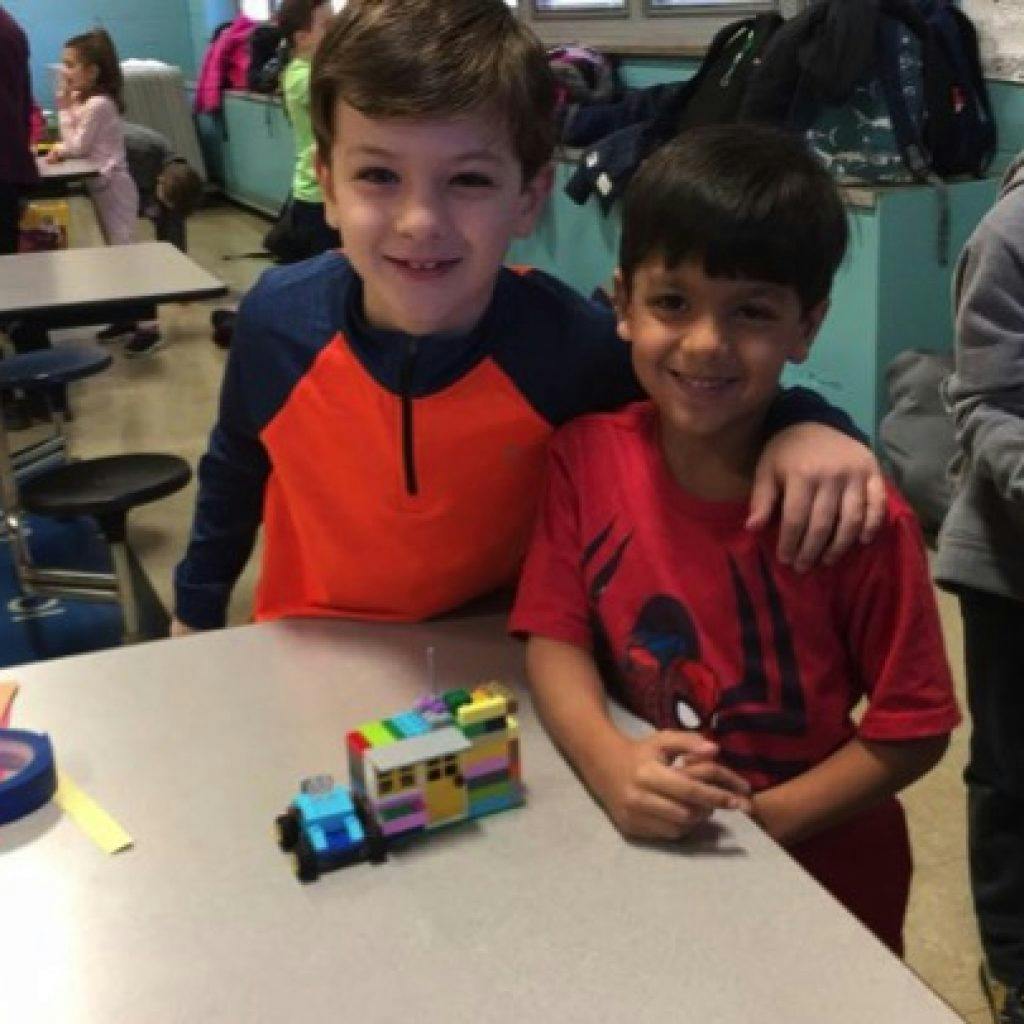 Two children standing next to a table with a toy made of building blocks