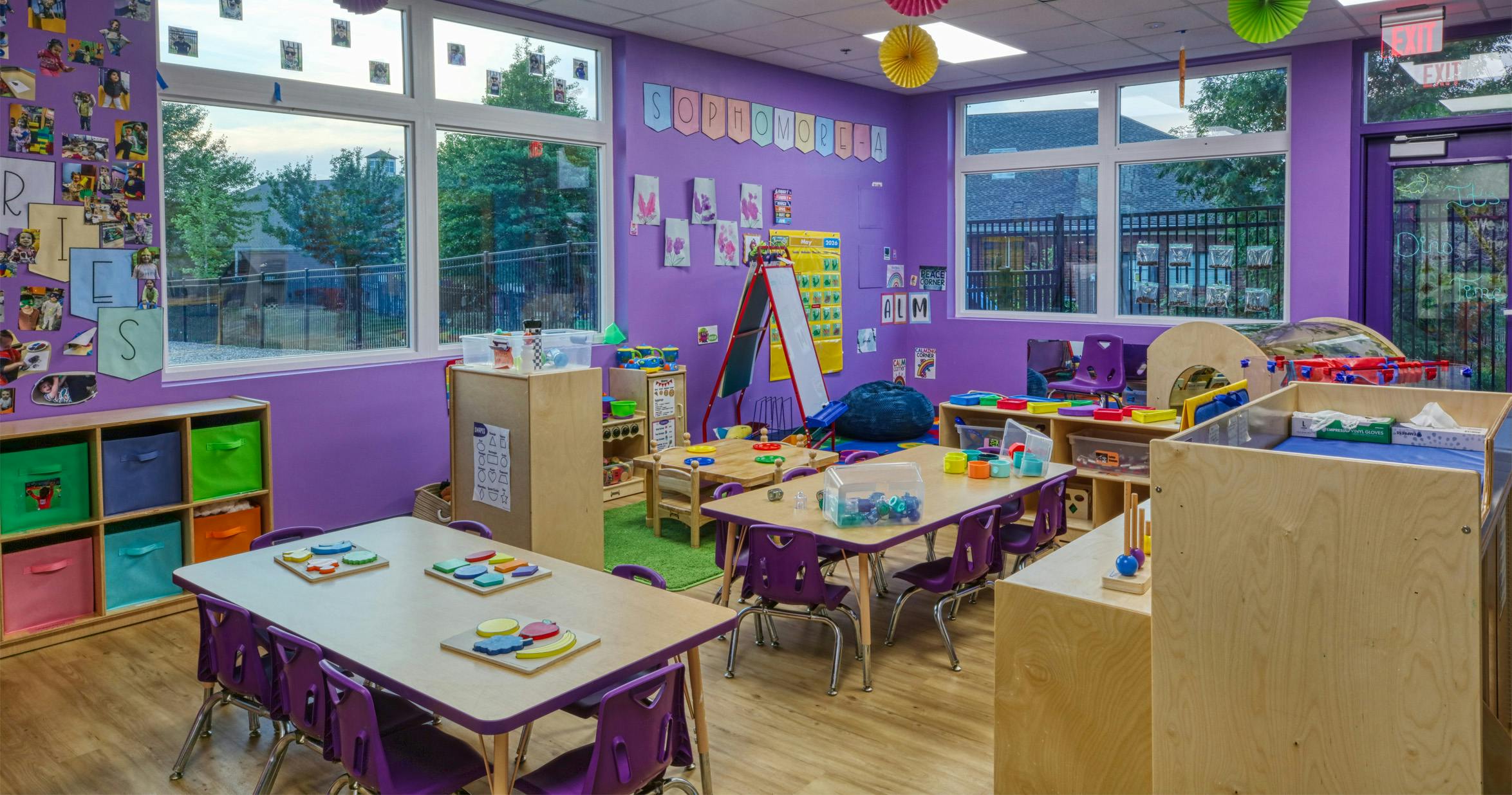 Colorful classroom with small tables and chairs, toys, and educational posters on purple walls