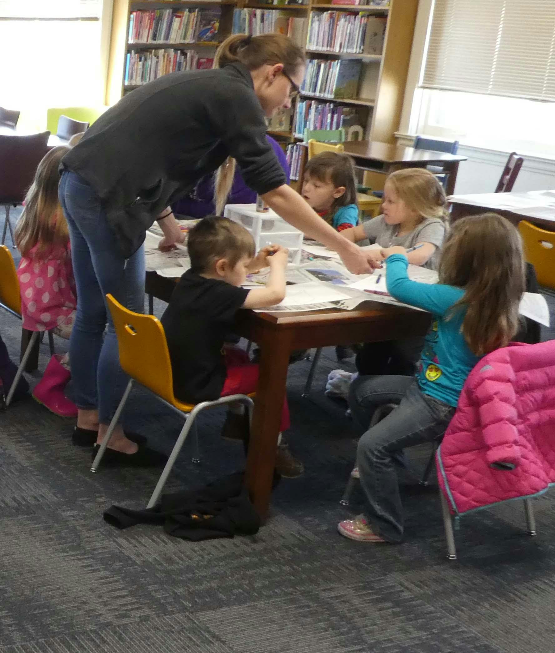 Person helping children at a table in a room with bookshelves