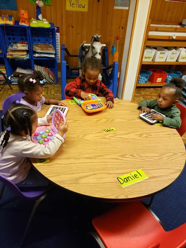 Four children playing with toy devices at a round table in a classroom