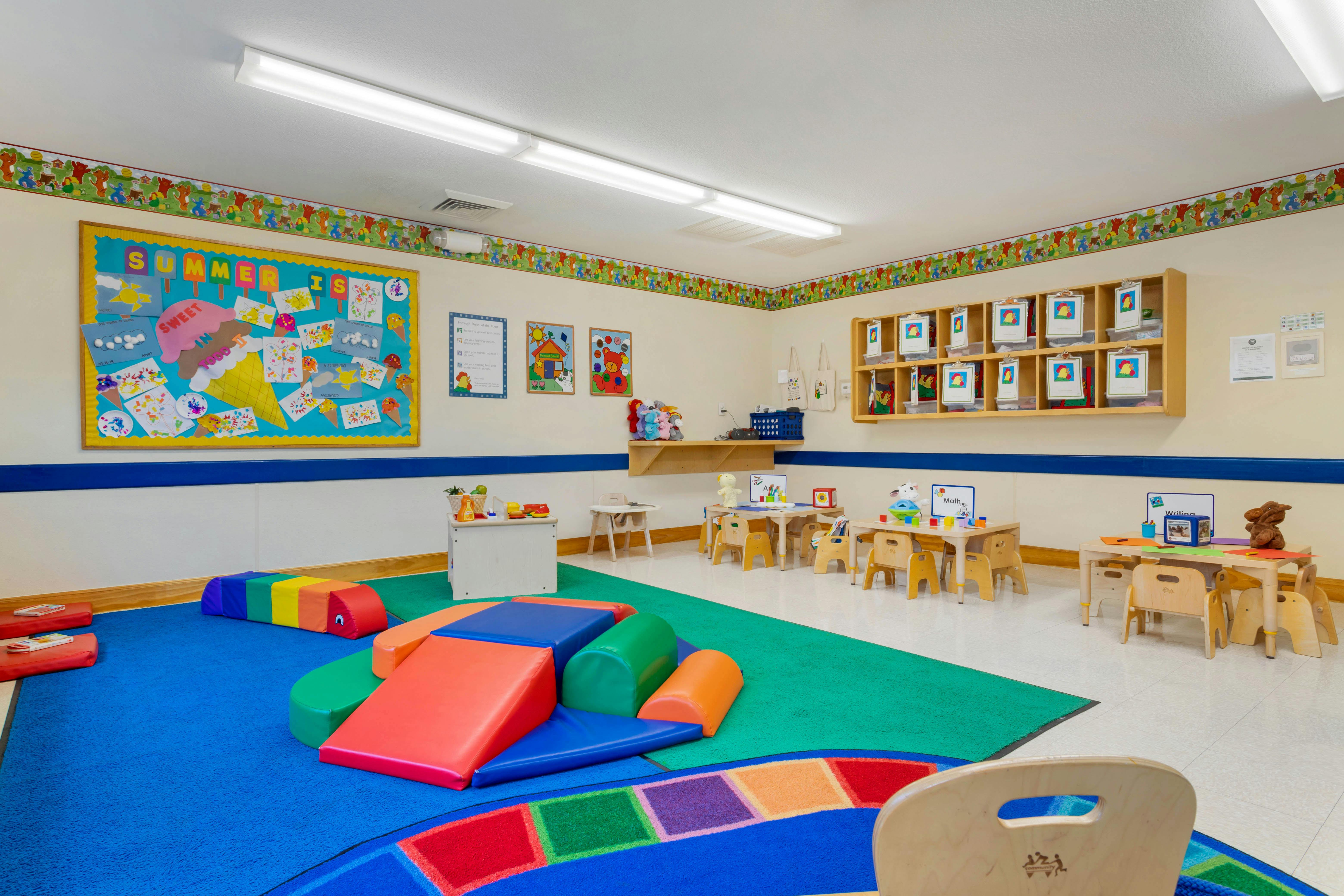 Children's classroom with small tables and chairs, soft play structures, and a bulletin board