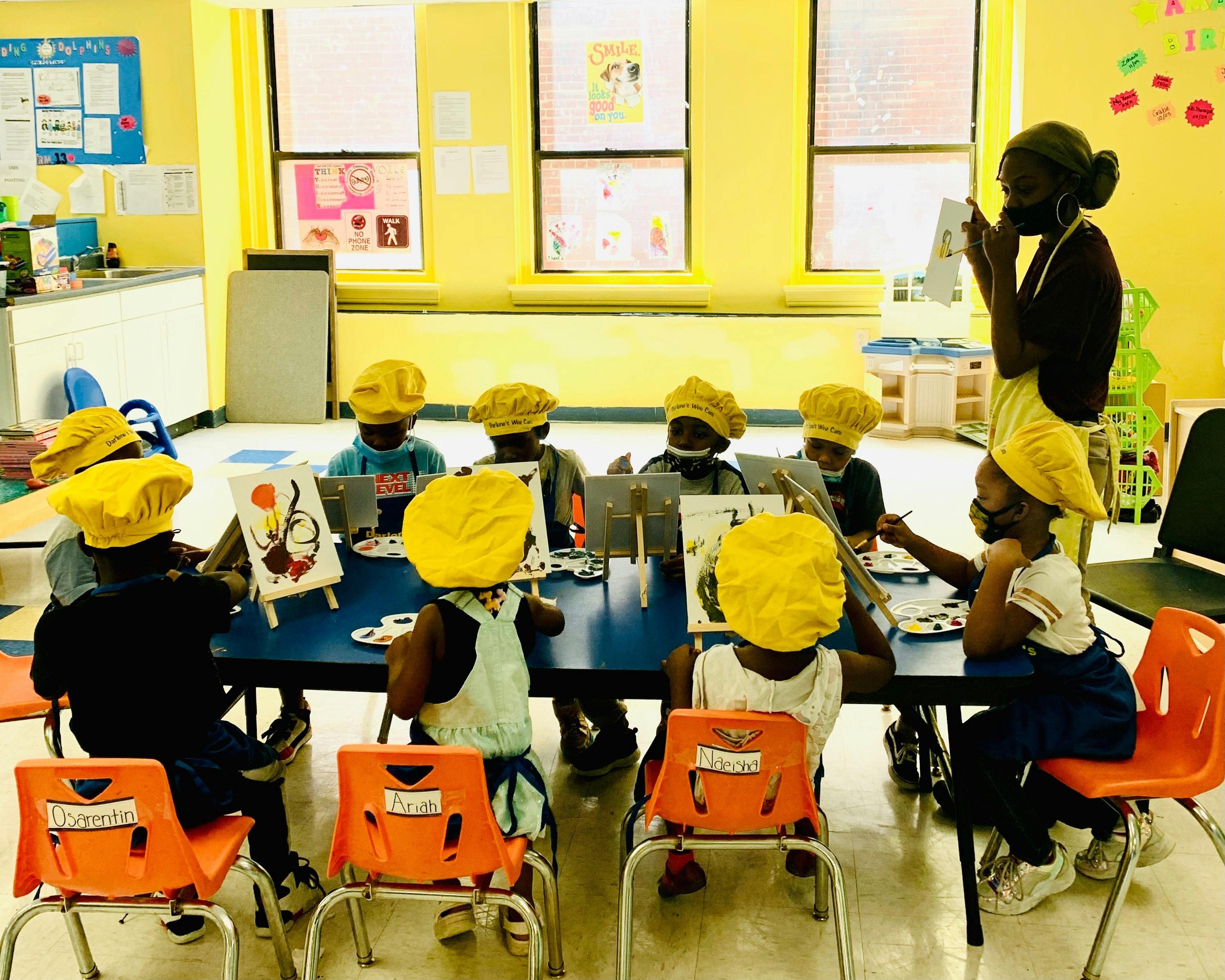 Adult and children in yellow hats painting at tables in a classroom