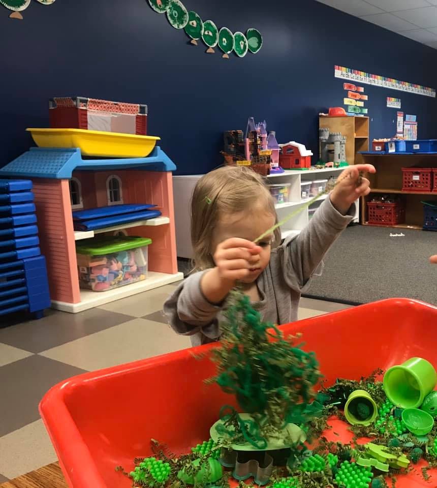 Child playing with green objects in a red bin in a classroom